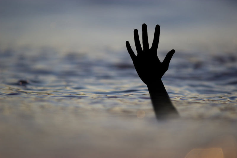 A silhouetted hand reaches out from beneath the water’s surface, frozen in a desperate grasp. The rippling water reflects the fading light, evoking a sense of loss and mystery. This image accompanies 'Return to the Water’s Edge: Austin’s Ongoing Haunting,