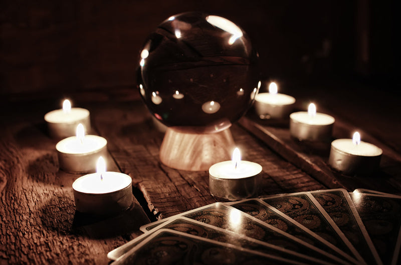 An ominous setting with a crystal ball placed on a rustic wooden table surrounded by flickering tea light candles and tarot cards. The dim light casts shadows that dance across the scene, evoking a sense of mystery and anticipation. Associated with Candle