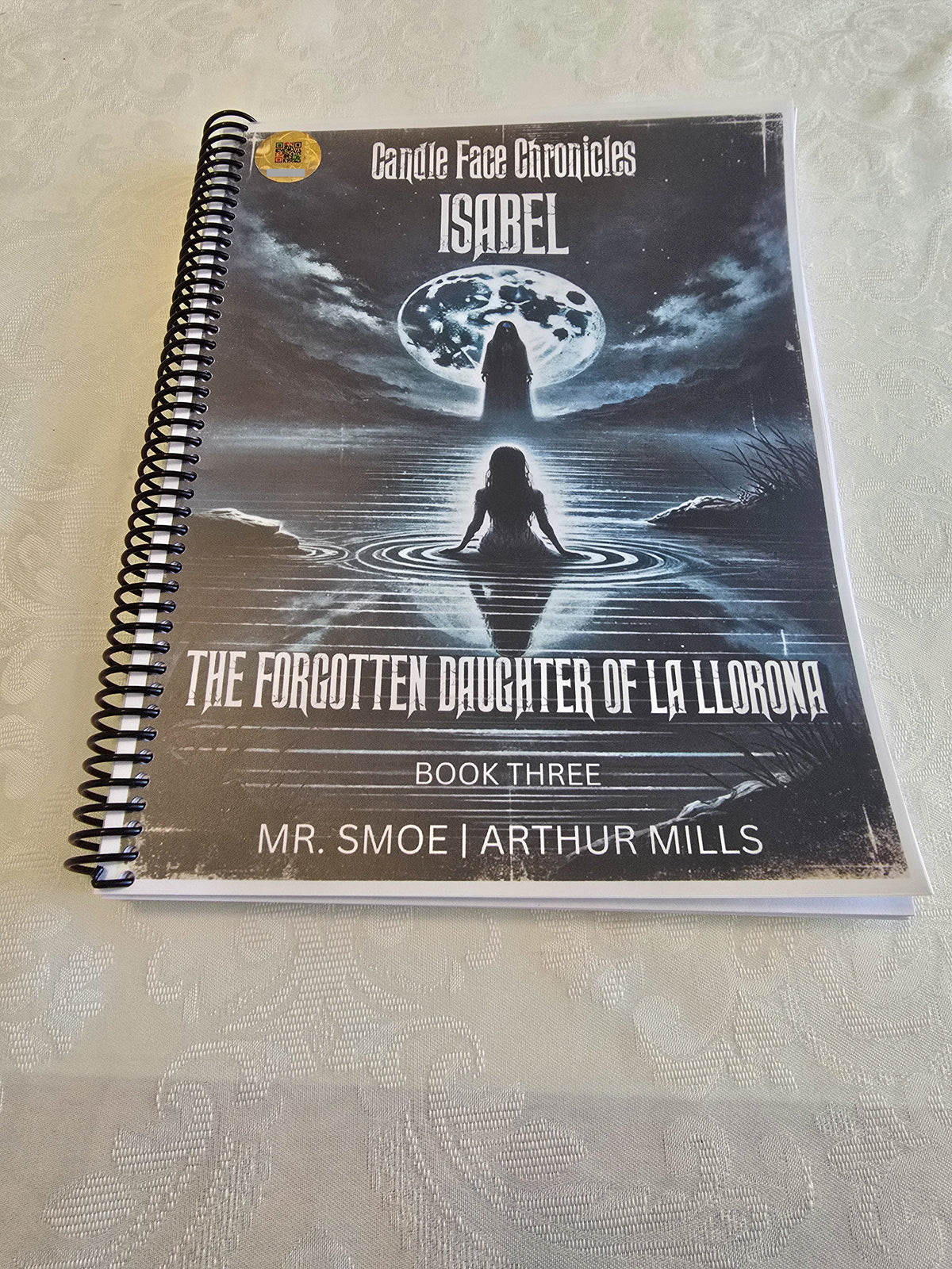 A spiral-bound copy of Candle Face Chronicles: Isabel – The Forgotten Daughter of La Llorona [Book Three] by Mr. Smoe and Arthur Mills. The cover art features a ghostly La Llorona standing before a glowing full moon while a young girl, Isabel, sits in the water below, symbolizing her tragic past and lost identity.
